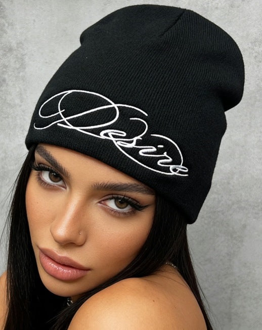 Black cuffless knit beanie with white embroidered script lettering, worn slouchy.