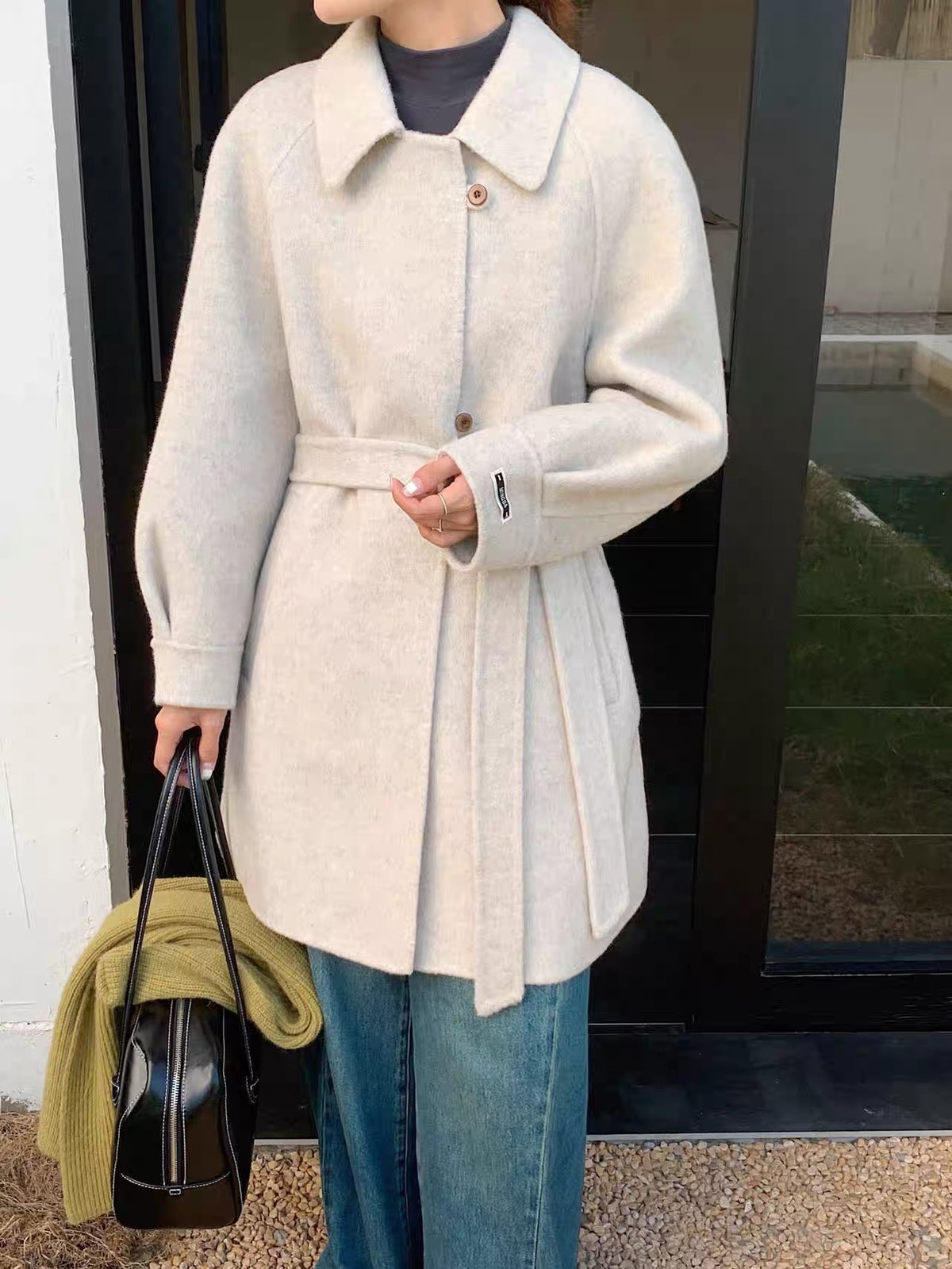 Off-white belted wool coat with raglan sleeves and single-breasted button closure, worn with jeans