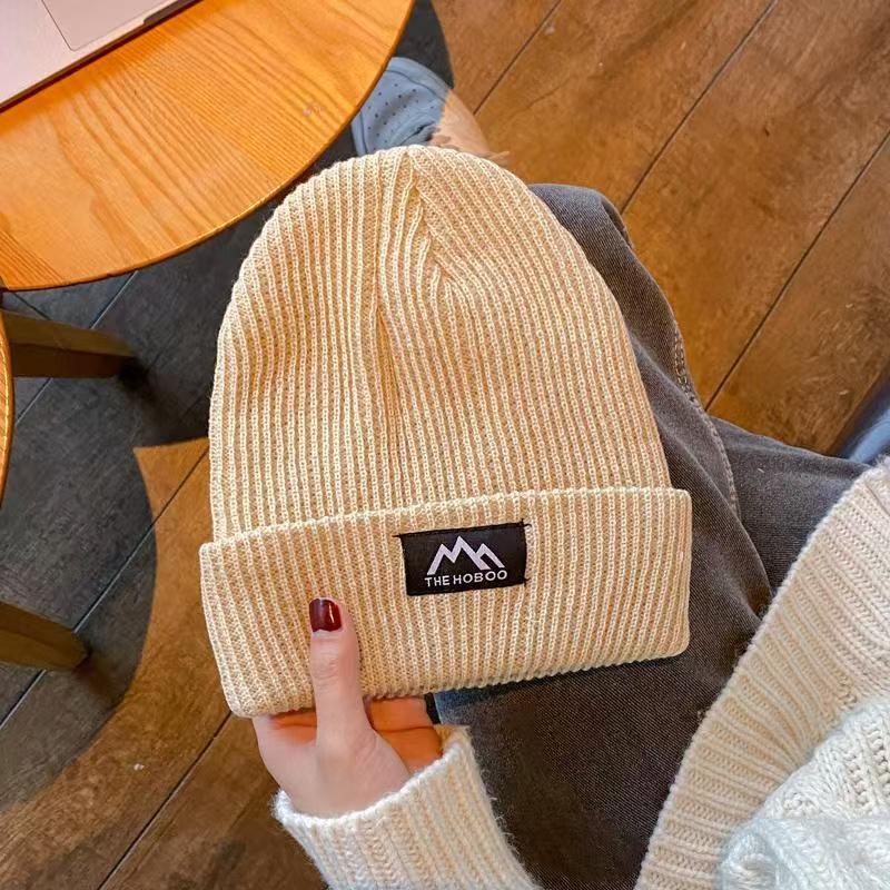 Beige ribbed knit beanie with folded cuff and woven brand patch, held by hand.